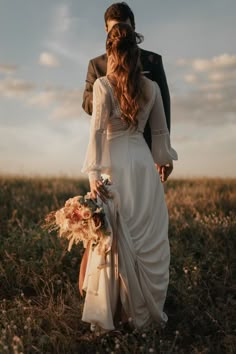 a bride and groom are walking through the grass together in an open field at sunset