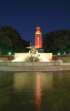 there is a clock tower in the distance behind a fountain with lights on at night