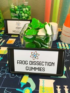 a table topped with lots of green gummy candies next to a sign that says frog dissection gummies