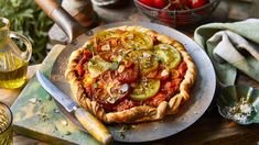 a pizza with tomatoes and other vegetables on a table next to olives, garlic, breadcrumbs and oil