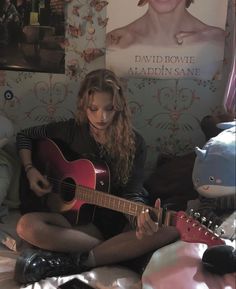 a woman sitting on top of a bed with a guitar in her hand and a poster behind her