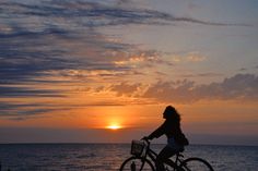 a woman is riding her bike on the beach at sunset with the sun in the distance