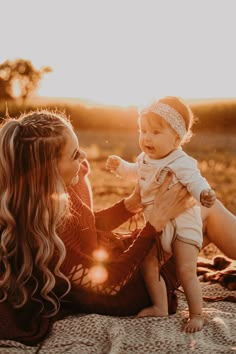 a woman holding a baby in her arms while sitting on a blanket with the sun behind her