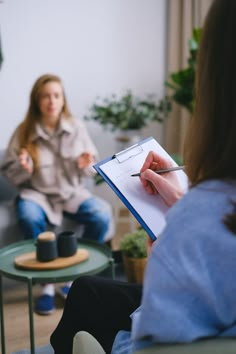 two women sitting on couches in a living room, one is writing and the other is holding a clipboard