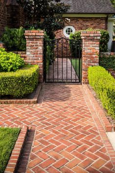 an iron gate in the middle of a brick walkway leading to a house with hedges and bushes