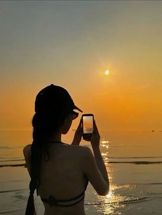 a woman standing on the beach taking a photo with her cell phone at sunset or dawn