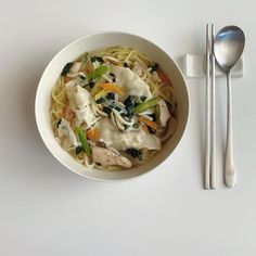 a bowl of noodles with chicken and vegetables next to a spoon on a white surface