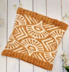 an orange and white knitted square sitting on top of a wooden table next to flowers