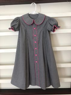 This child's size 6 dress was made with cotton gingham and it has 7 pink snaps on the front. It has a pleat on either side of the front band and two pleats in the back. It has a round collar and puffed sleeves with elastic that gathers the sleeve. There is pink mini-piping down the center band, around the collar, and on the edges of the sleeves. It measures 26' long from the highest point of the shoulder to the bottom of the hem. I can shorten this dress if needed. It can be washed in the washing machine and dried in the dryer. Collared Spring Dresses For School, Spring School Dress With Collar, Spring School Dresses With Buttons, Fitted Black Dress For School, Black Fitted Dress For School, Cute Fitted Dress With Button Closure, Pleated School Dress For Spring, Spring Pleated School Dress, Cute Fitted Pleated Dress