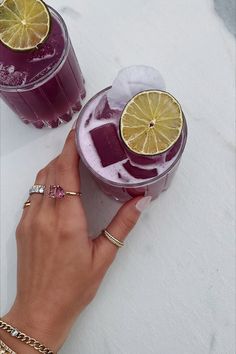 a woman holding two glasses filled with purple liquid and topped with lemon slices, on top of snow covered ground