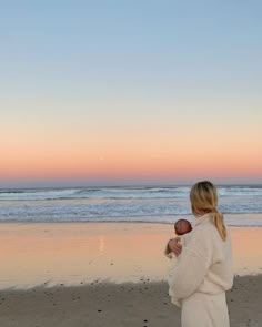 a woman standing on top of a sandy beach holding a baby in her arms and looking at the ocean