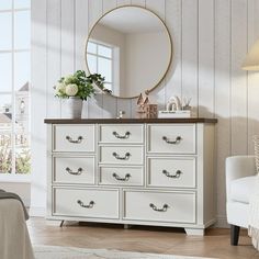 a white dresser sitting next to a bed in a bedroom under a large round mirror