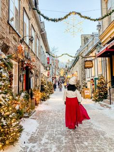 a woman walking down a snowy street with christmas decorations on the buildings and trees around her