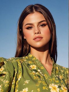 a woman with long brown hair wearing a green floral shirt and gold earrings, standing in front of a blue sky