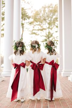 three girls in white dresses with red sashes and flower crowns on their heads are looking at each other