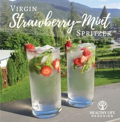 two glasses filled with ice and strawberries on top of a table next to trees