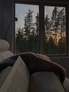 an open book sitting on top of a bed next to a window with trees outside