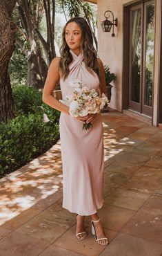 a woman in a pink dress standing on a patio holding a bouquet and looking at the camera