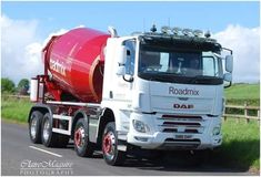 a red and white truck driving down a road next to a lush green field on a sunny day