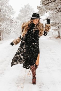 a woman walking down a snow covered road wearing a black dress and hat with fur