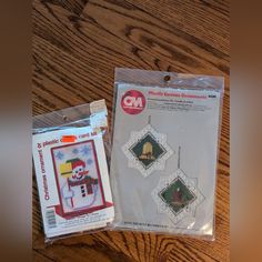 two cross - stitch christmas ornaments in plastic bags on a wooden table next to each other