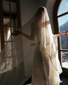 a woman in a white dress standing next to a window with a veil over her head