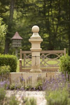 a stone fountain in the middle of a garden with lavenders and trees around it