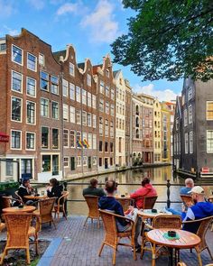 people sitting at tables on the side of a river in front of some old buildings