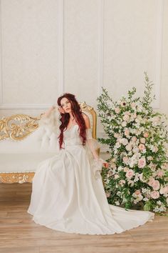 a woman in a white dress sitting on a gold bench next to a flower arrangement