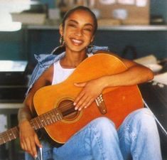 a woman sitting in a chair holding an acoustic guitar and smiling at the camera with her arms crossed