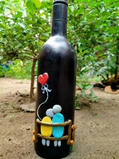 a black bottle with balloons painted on it sitting in the dirt near some trees and bushes