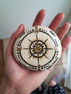 a hand holding a rock with a compass painted on it's side, in front of a tile floor