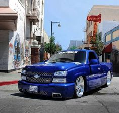 a blue truck parked on the side of a road next to tall buildings and shops