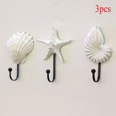 three seashells hang from hooks on a white wall, one is holding a starfish and the other has a black hook