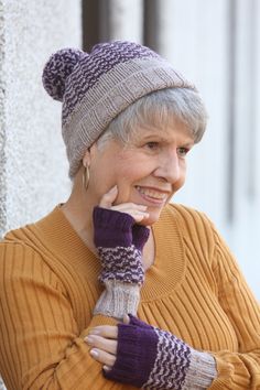 an older woman with grey hair wearing a knitted hat and mittens, leaning against a wall