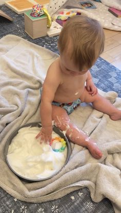 a toddler sitting on the floor playing with some white frosting in a bowl