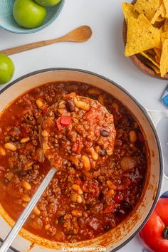 a bowl filled with chili and beans next to some chips, limes and tomatoes