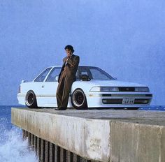 a man standing next to a white car on top of a pier near the ocean