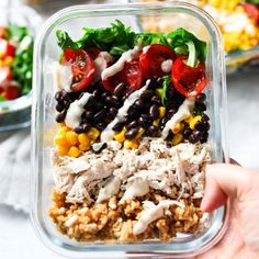 a person holding up a plastic container filled with salad and rice, including tomatoes, black beans, corn, lettuce