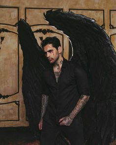 a man with tattoos standing in front of a black angel wings sculpture and holding his hands on his hips