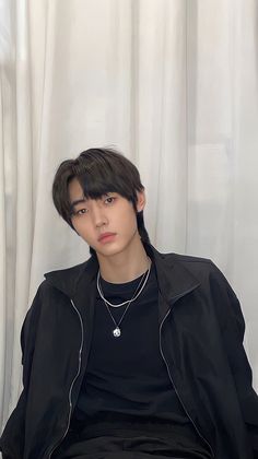 a young man sitting in front of a curtain wearing a black shirt and silver necklace