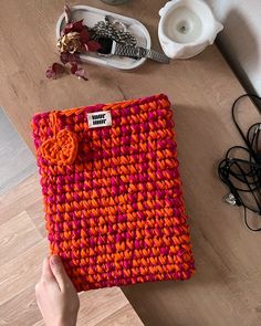 a hand holding an orange and pink knitted case on top of a wooden table