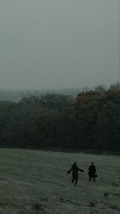 two people running through a field with a kite flying in the sky over them and trees behind them