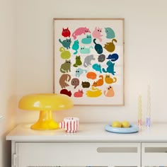 a white dresser topped with a yellow lamp next to a wall mounted art print on the wall