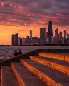 two people are sitting on steps overlooking the water with city skyline in the background at sunset