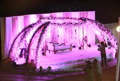 people standing in front of a stage decorated with purple flowers and white draping