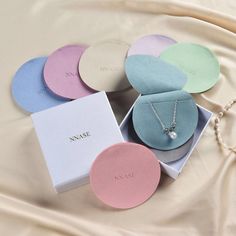 six round boxes with personalized necklaces in them on a white satin surface next to a pearl necklace