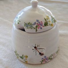 a ceramic jar with a bee painted on the side and flowers around it, sitting on a white tablecloth