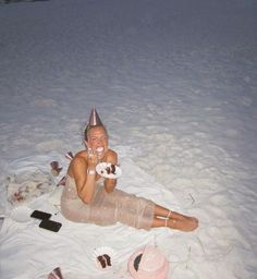 a woman is sitting in the snow eating cake and drinking icecream while wearing a party hat