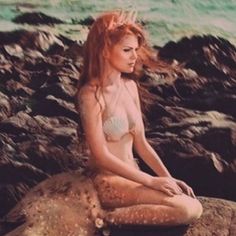 a woman sitting on top of a rock next to the ocean with her hair blowing in the wind